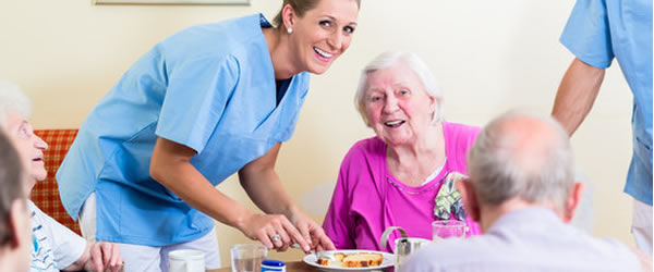 Nurse with a senior woman at a table with a plate of food in front of them.  Other seniors sitting around the table not in focus.